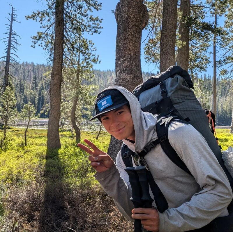 A person wearing a gray hooded sweatshirt, a black baseball cap, and a large backpack is standing in a forest. They are holding trekking poles and making a peace sign with their left hand. The background includes trees, green grass, and a body of water in the distance. The sky is blue and clear.